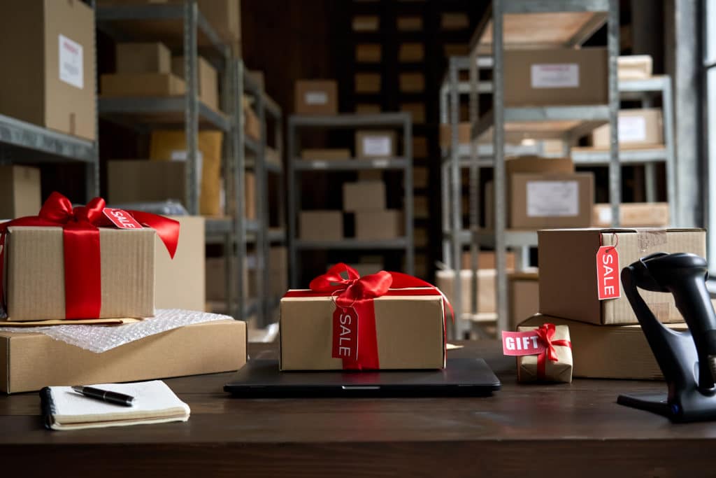A logistics warehouse with boxes on shelves. A few boxes on a desk wrapped in red ribbon with tags that read "sale" and "gift" with a laptop and notepad for shipping.