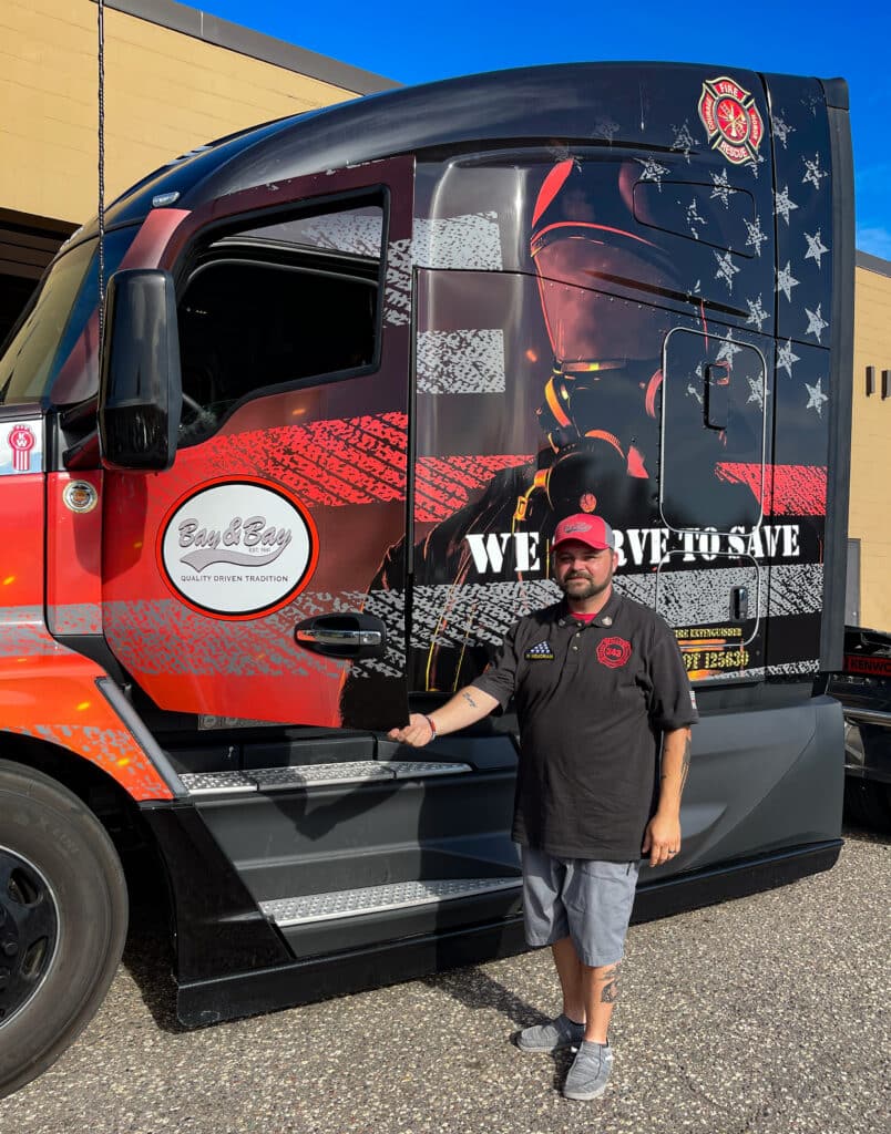 A photo of a Bay & Bay driver standing outside of the passenger door of his first responder wrapped Bay & Bay Kenworth truck. There is a firefighter on the side of the truck with text that reads 'We Serve to Save.'