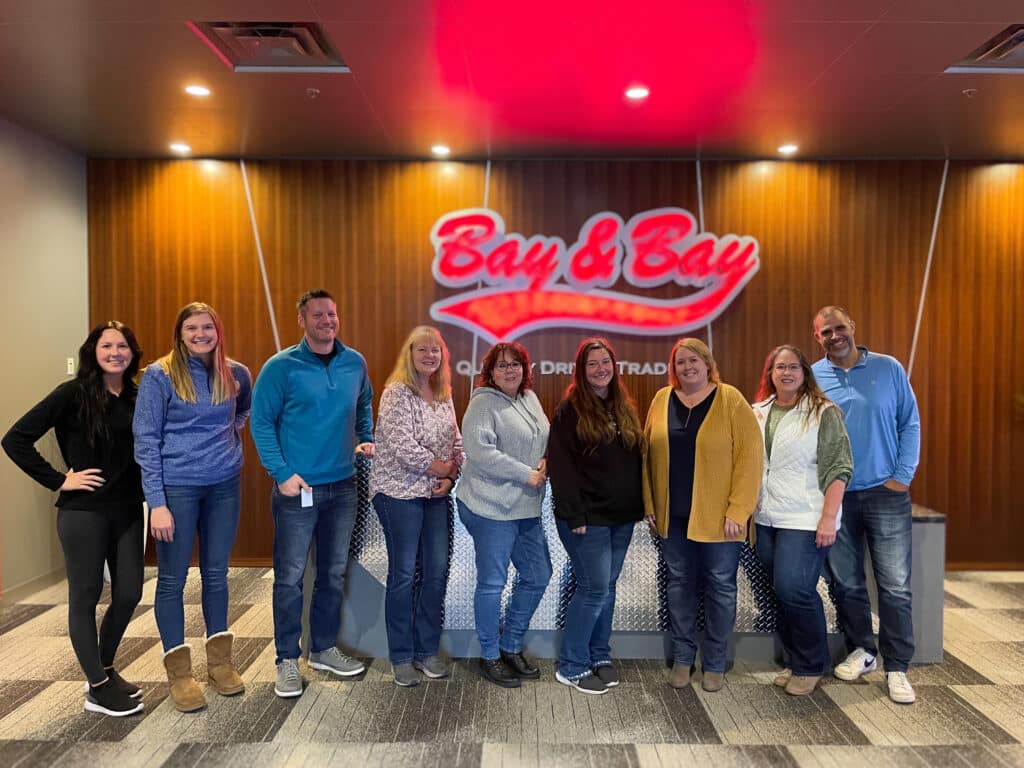 A photo of Bay & Bay's accounting team at the Eagan, MN corporate office. They are smiling in the main lobby with the Bay & Bay logo in the background.