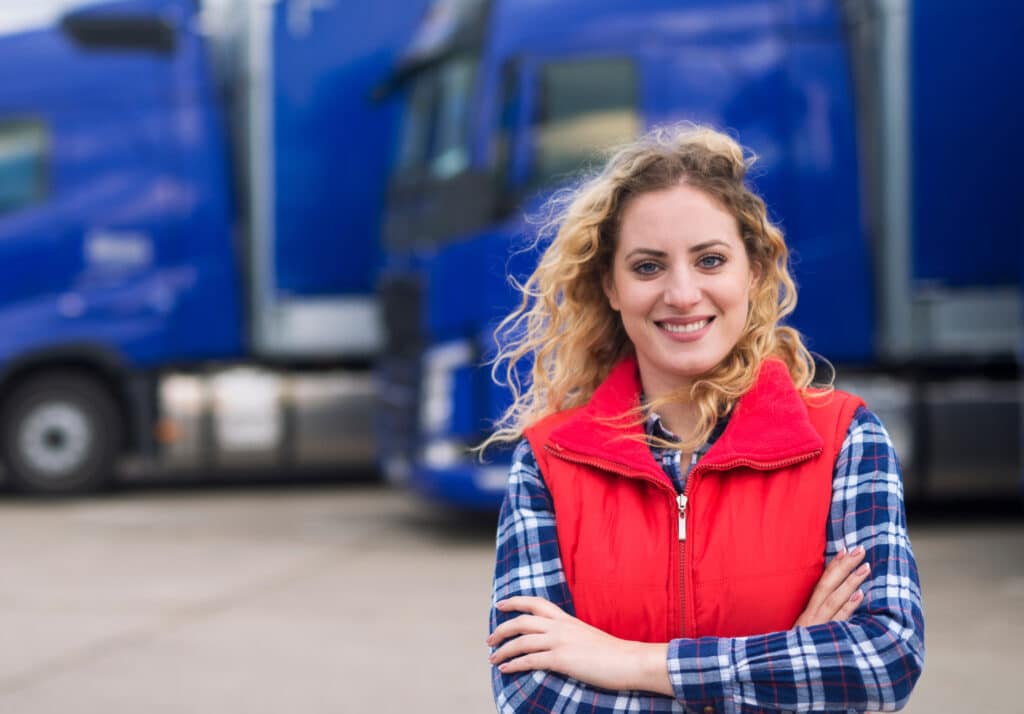 A photo of a female truck driver smiling with her arms crossed in front of a line of blue semi-trucks. She is representing women in the trucking and transportation industry.