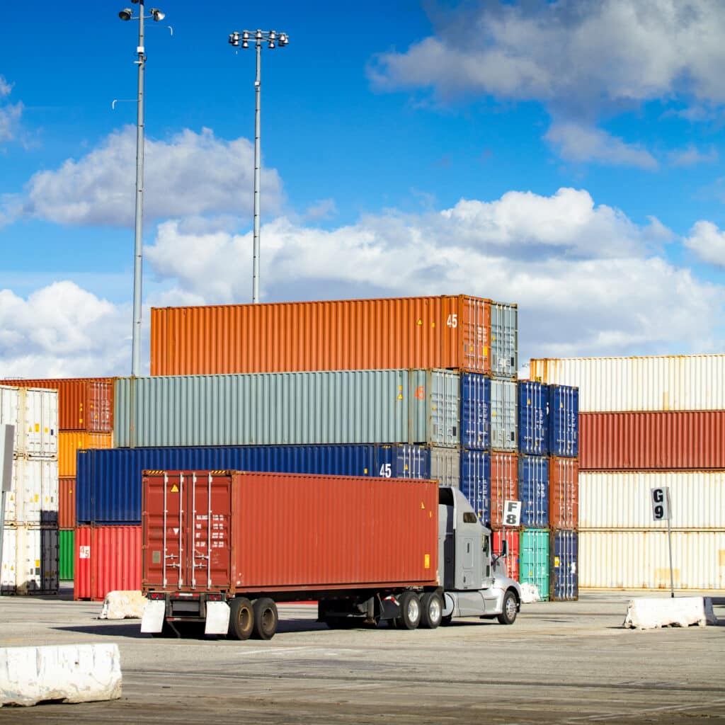 A photo of many intermodal shipping containers at a warehouse. One red shipping container is loaded onto a truck ready to ship to the customer.