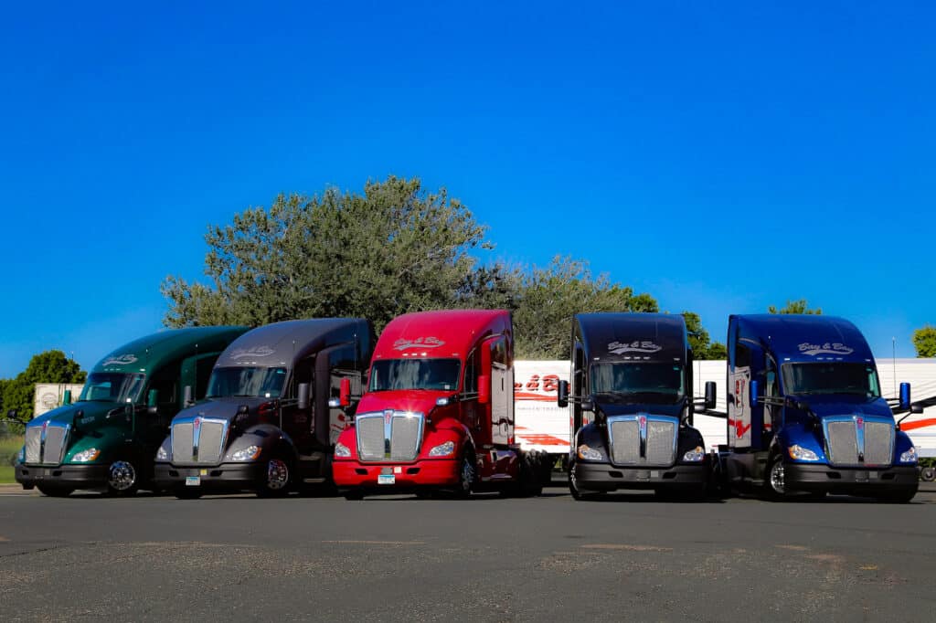 A photo of 5 Bay & Bay Kenworth T680 trucks in front of two Bay & Bay refrigerated trailers. The trucks are green, grey, red, black, and blue.