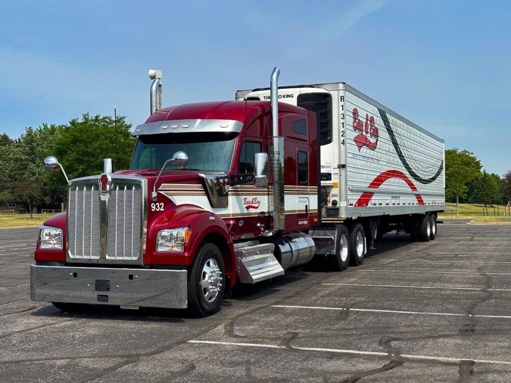 A photo of a new Bay & Bay Kenworth W990 that is red with white and gold stripes. A Bay & Bay refrigerated trailer that has red and black stripes, and the Bay & Bay logo is on the side of the truck.
