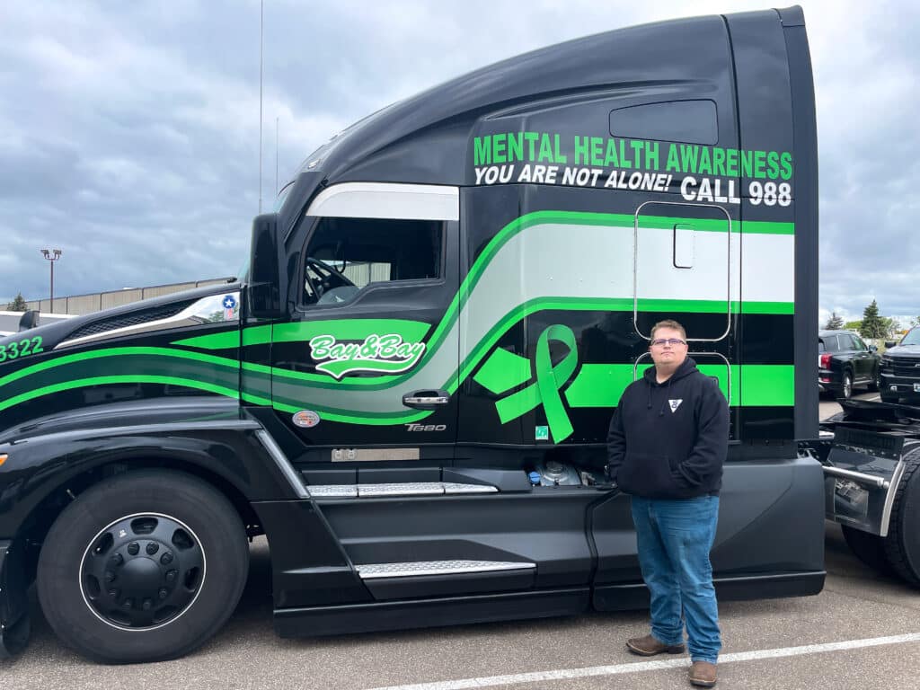 A photo of Bay & Bay Owner Operator, Greg Ekstrom standing in front of his black and green Kenworth T680 truck with a mental health awareness wrap.