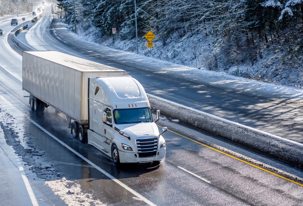A photo of a white truck driving down a road during the winter with snow. This driver has their winter trucking essentials to ensure a safe drive to his destination.