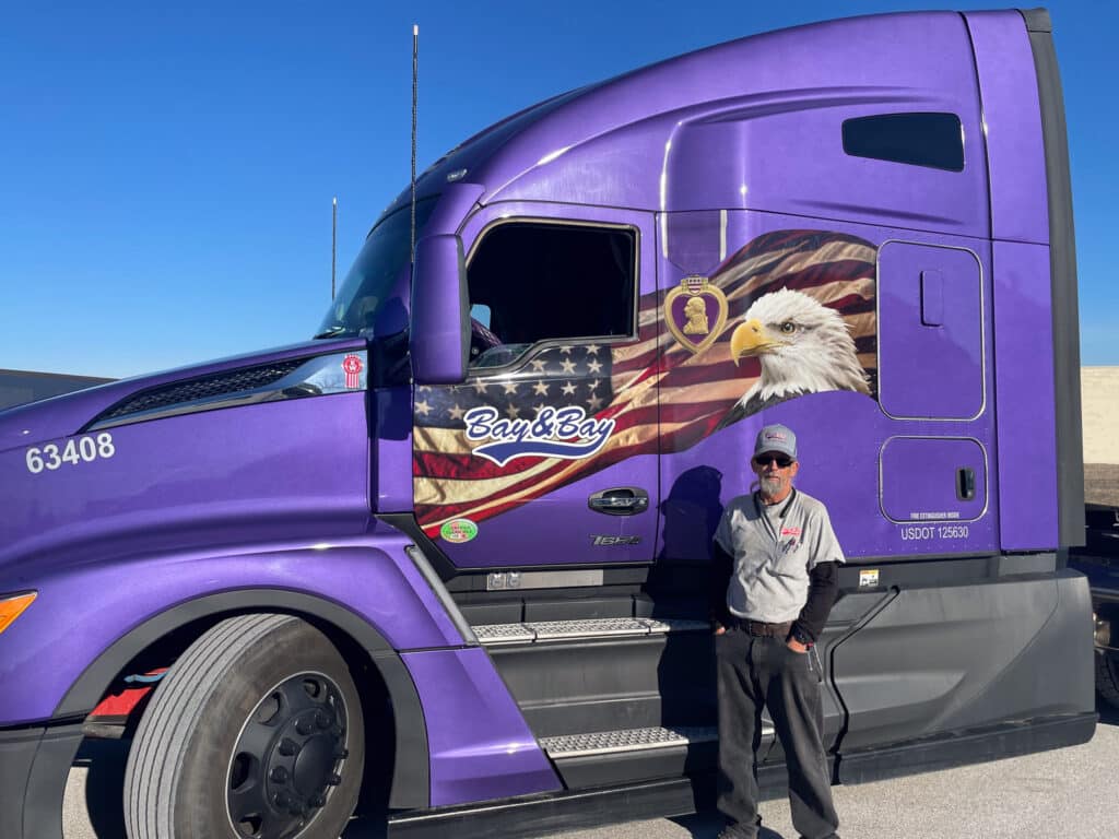 A photo of a purple Bay & bay Kenworth T680 truck, with owner operator Jamie Gardner standing in front of it. The purple truck is wrapped in an Army Purple Heart theme to honor those that didn't receive their Purple Heart.
