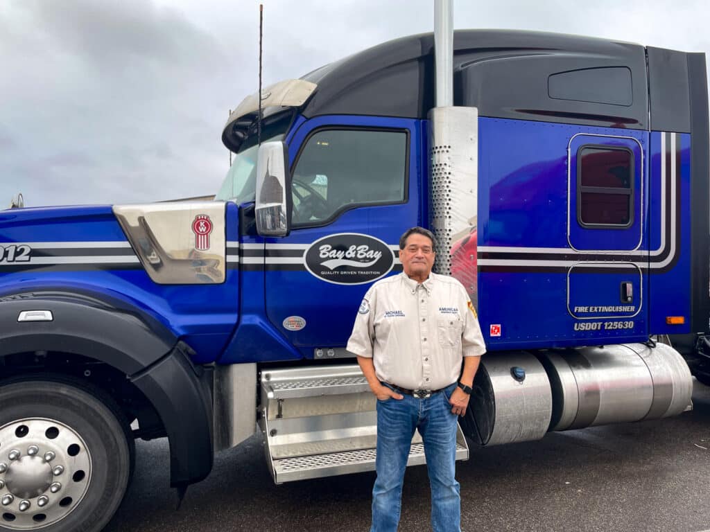 Bay & Bay Lease Purchase Driver, Michael Udasco standing in front of his excellent blue and silver W990, wearing a shirt that says 50 years of excellence.